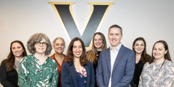 A group of about 10 genetic counselors, mostly women in business attire, pose for a photo in front of the Vanderbilt V logo.