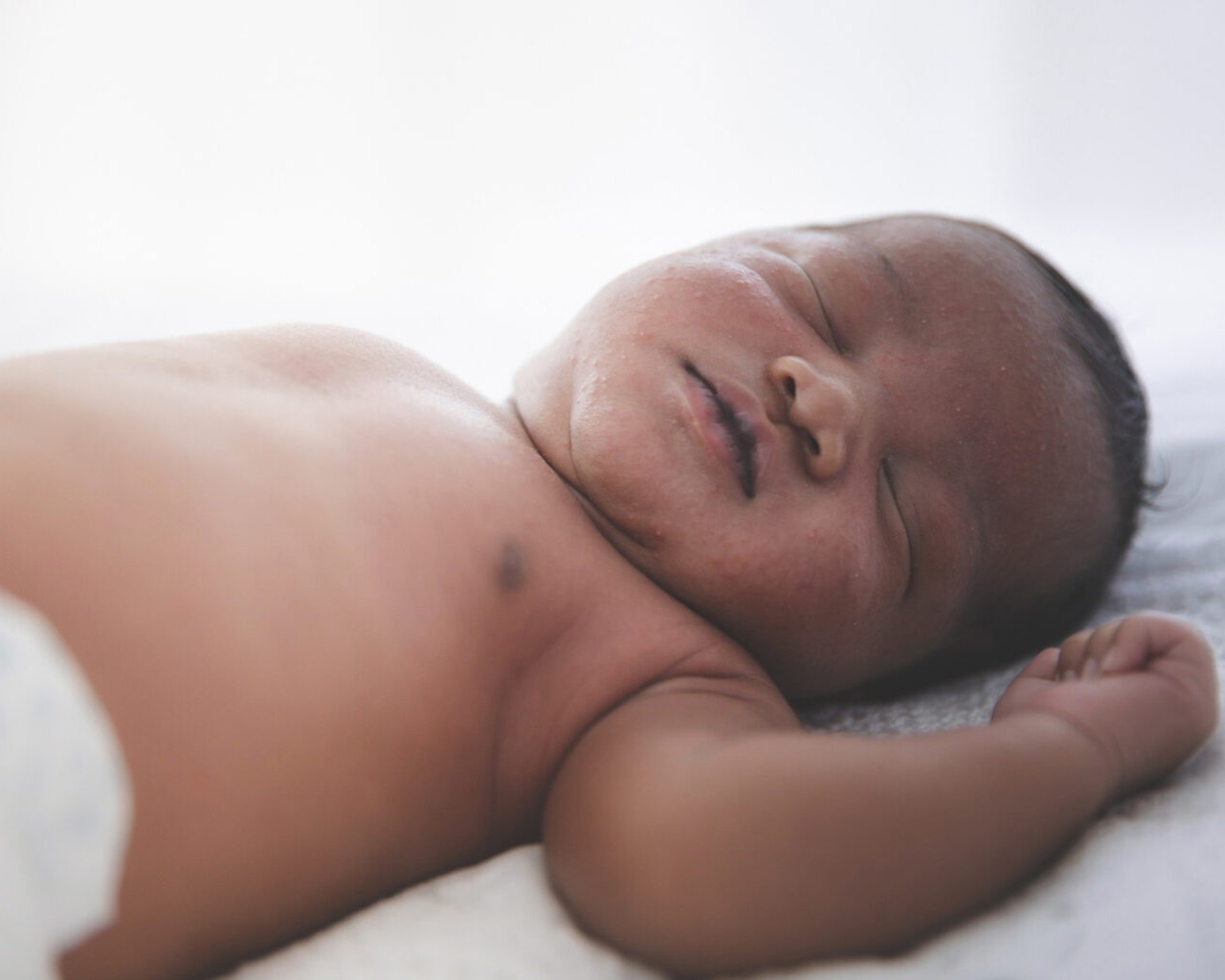 An African American baby in a diaper sleeps peacefully on a white sheet.