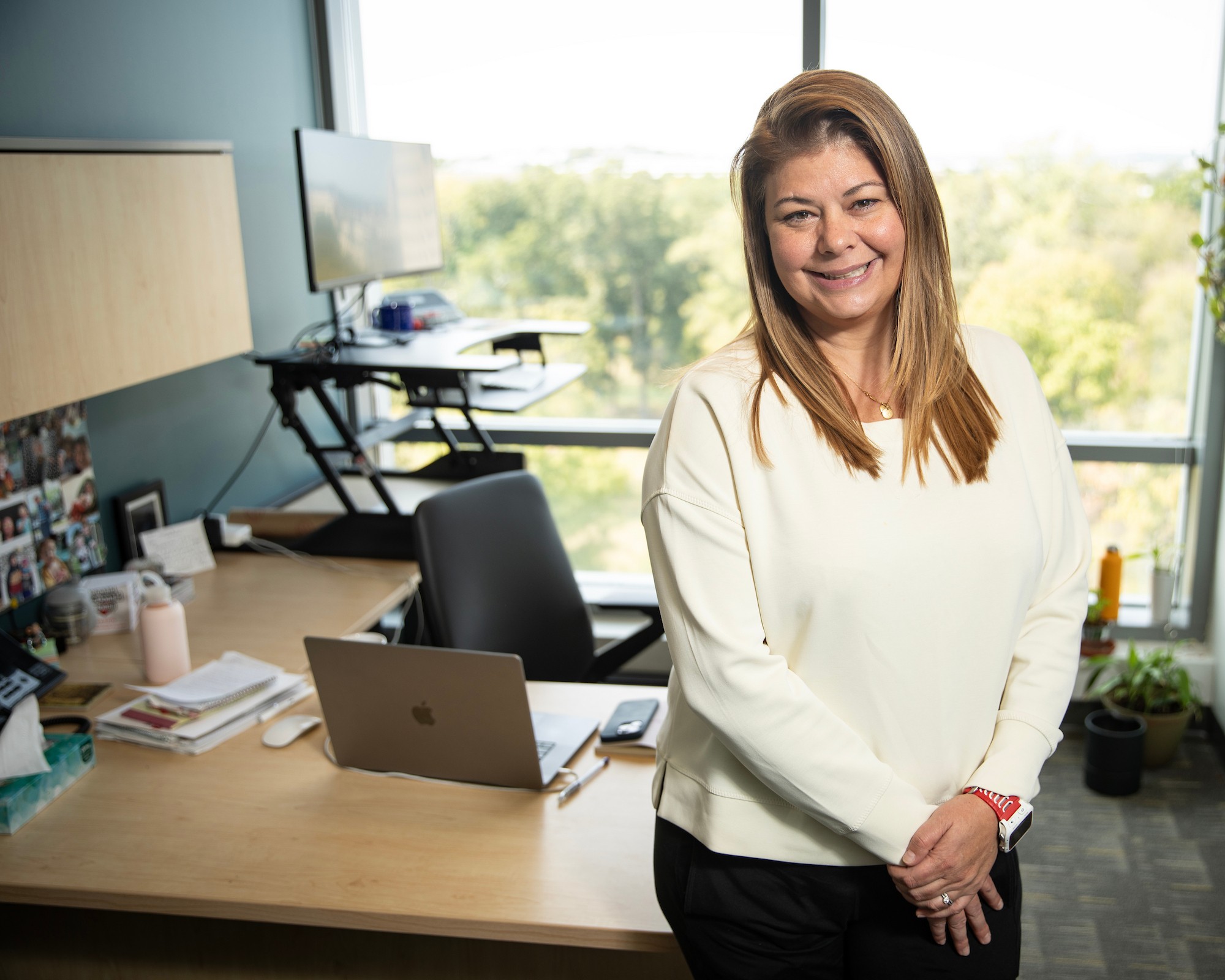 Dr. Piper Below, in a white top and black pants, poses in front of her desk with a view of trees through her window.
