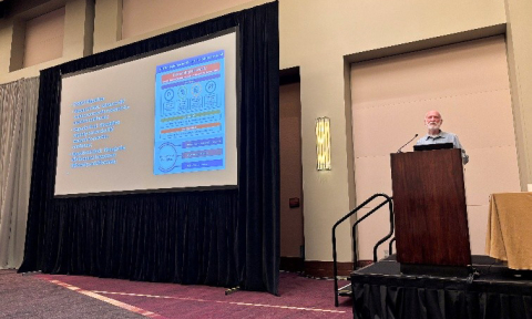 Dr. Kendall Bryant presents from a podium with a large screen to his left.