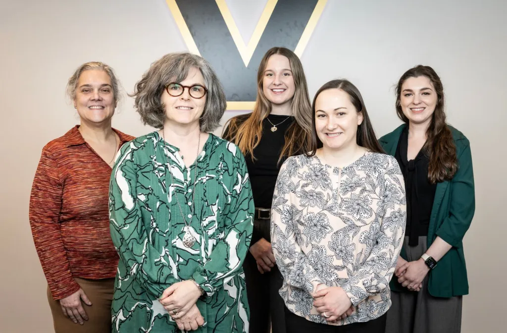 A group of five white women (genetic counselors) pose in front of a Vanderbilt V logo.