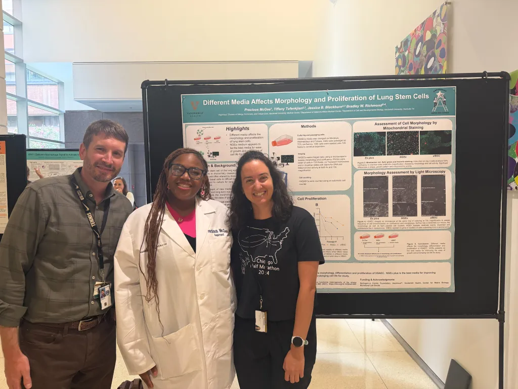 One male and two female researchers pose in front of a scientific poster during a conference.